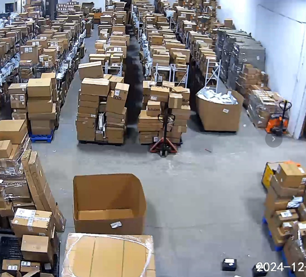 Aerial security camera view of R3 Goods Flow warehouse in December 2024 (2024-12) showing organized inventory rows with worker and pallet jacks